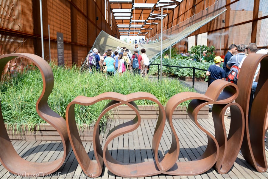 A brown metal sign of Brazil in front of grass and a suspended cargo net at Expo 2015